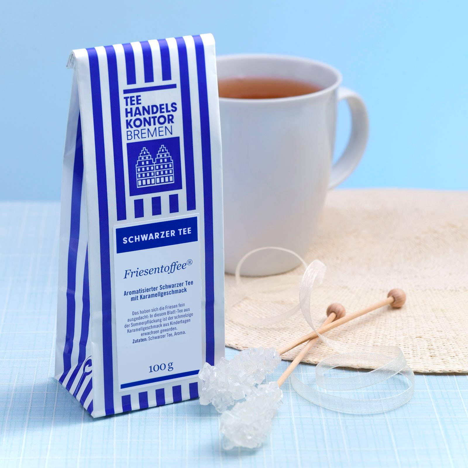 Dekoratives Bild: aufgebrühter Schwarztee in weißem Mug mit blau-weißer Teetüte und Kandissticks