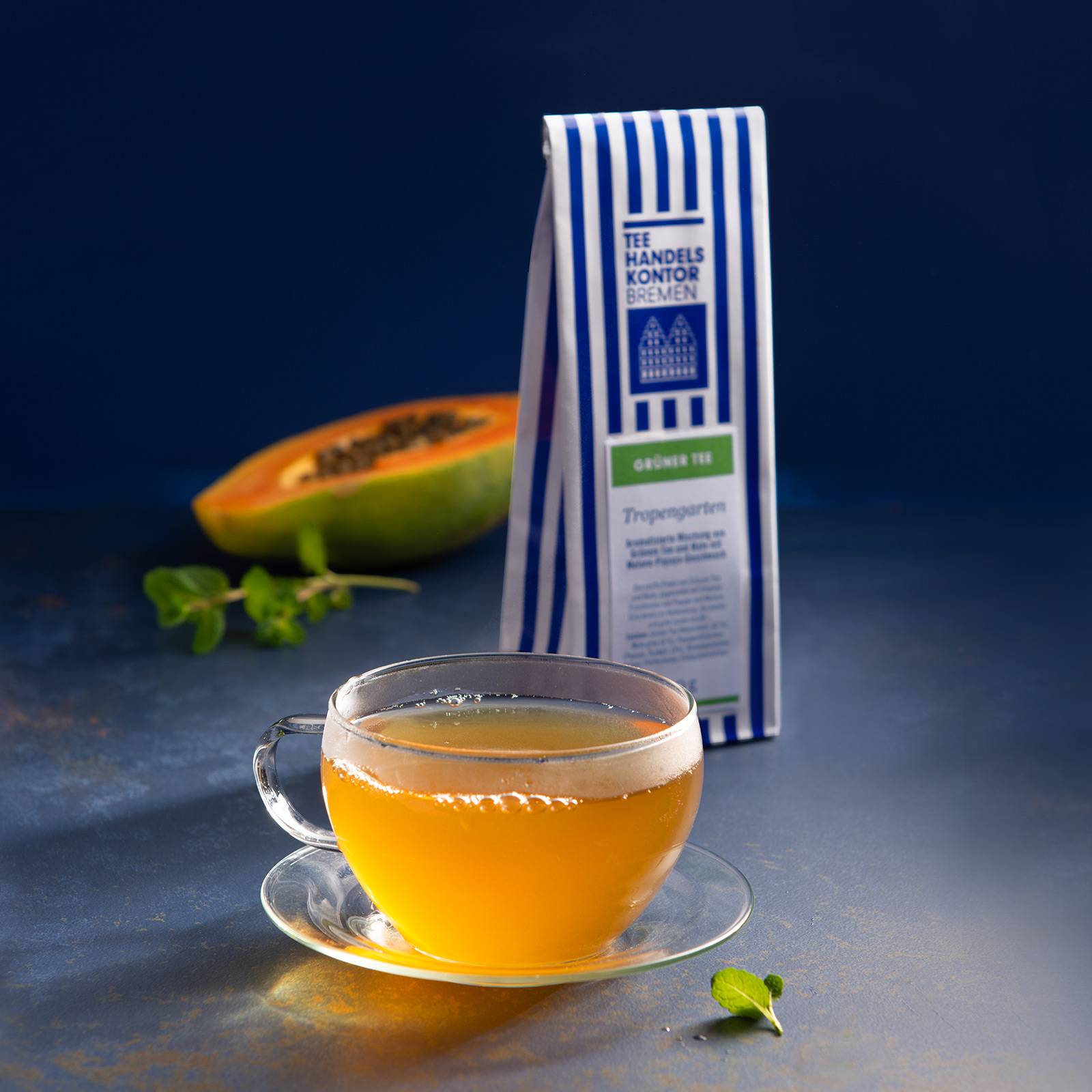 Dekobild: 1 Glasteetasse gefüllt mit Tee, daneben eine blau-weiß gestreifte Teetüte und eine Papaya