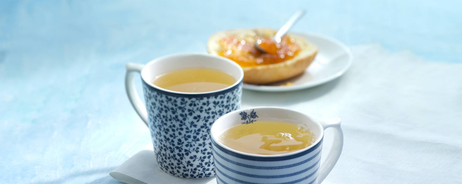 2 Porzellanbecher mit Kräutertee sowie ein mit Konfitüre bestrichenes Brötchen